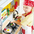 A child wearing a red chef hat and apron plays with the HAPE Deluxe Kitchen with Fan Fryer. They are holding a can above a toy pot on an electric stove and have a toy fried egg in a pan. The HAPE kitchen playset features various accessories, including utensils, a pasta dish, and condiment bottles, perfect for imitative play.