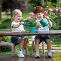Two children, a boy and a girl, sit on small stools at a wooden table outdoors, smiling as they use green watering cans from their HAPE Garden Tool Set by HAPE to water a potted plant. The lush garden background features blooming flowers and greenery, accented with charming plant signs.