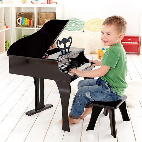A young boy in a green shirt and jeans sits on a small bench, playing the HAPE Happy Grand Piano, Black in a bright, colorful room with various toys and books on shelves in the background. He is smiling and appears engaged with the piano.