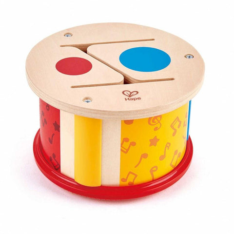 The Hape Double-Sided Hand Drum features a wooden tone drum with a red and yellow base adorned with musical notes and instruments. The drumhead displays both a blue circle and a red circle, complete with small wooden mallets attached. Create wild rhythms with this versatile drum, which proudly displays the Hape logo on top.