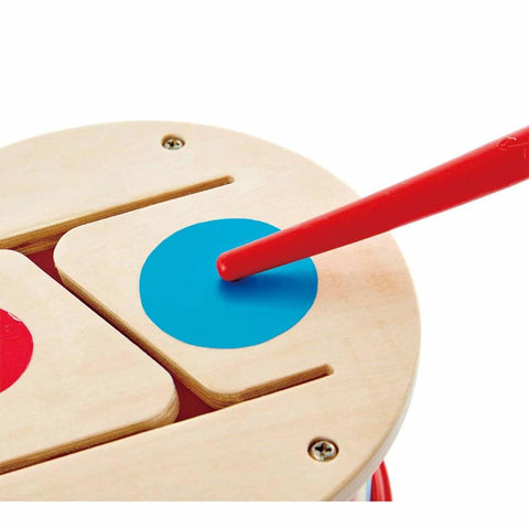 Close-up of a Hape Double-Sided Hand Drum with a red drumstick striking a blue dot on one of the drum pads. This versatile drum by Hape features a natural wood finish and clean, simple design, perfect for creating wild rhythms.