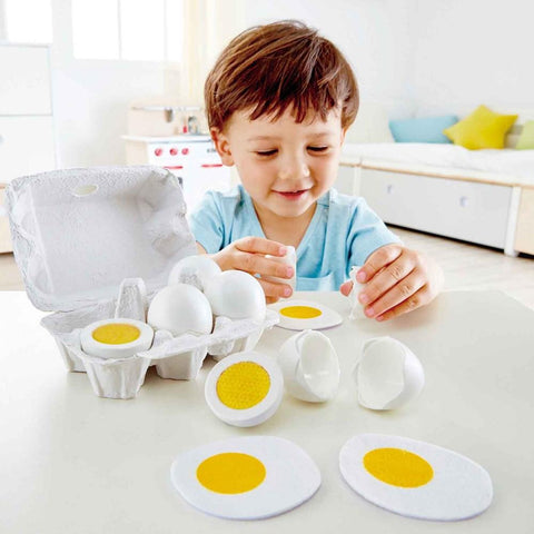 A young child is seated at a table, engaging in realistic play with the Hape Egg Carton set. The child is opening plastic eggs from the Hape brand, revealing halves that resemble hard-boiled eggs with yellow yolks. An egg carton containing additional toy eggs from Hape is also on the table.