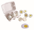 A Hape Egg Carton from Hape holds three white cardboard eggs, while beside it, four additional white plastic eggs are cracked open to show yellow centers. One of the cracked eggs reveals a yolk connected to a piece of white felt. The setup portrays toy eggs intended for realistic play, emulating fried eggs.