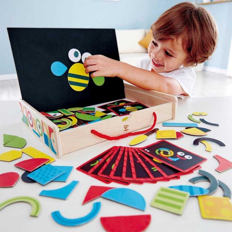 A smiling child plays with the Hape Magnetic Art Box, fostering creativity and engagement. The wooden set from Hape contains an array of colorful geometric shapes and pattern cards. The child places a shape on the magnetic board, creating a fun and stimulating learning activity.