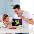 A man and a young girl are sitting at a table, smiling and looking at colorful puzzle pieces. The girl is placing magnetic shapes from the Hape Magnetic Art Box onto a small black board, creating a bird. The Hape Magnetic Art Box is also on the table. The man holds a piece and encourages the girl's creativity.