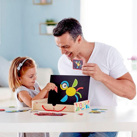 A man and a young girl are sitting at a table, smiling and looking at colorful puzzle pieces. The girl is placing magnetic shapes from the Hape Magnetic Art Box onto a small black board, creating a bird. The Hape Magnetic Art Box is also on the table. The man holds a piece and encourages the girl's creativity.