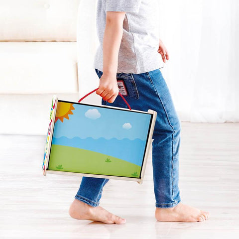 A child wearing blue jeans and a light gray shirt is walking indoors carrying the Hape Magnetic Art Box, a colorful wooden case with a landscape design on the side. This creative toy from Hape, featuring a red handle, fosters imaginative play. The room features light-colored hardwood flooring.