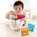 A young child plays with the Hape Mix & Bake Blender, turning the sturdy wooden crank to mimic mixing ingredients in a white bowl. The child's imagination comes alive, surrounded by toy packets of flour and sugar, each labeled and illustrated with pictures. The scene is set perfectly for a fun baking adventure in the background.