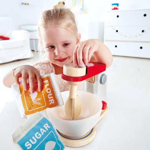 A young child with blonde hair is playing with the high-quality Hape Mix & Bake Blender from Hape. She is holding a packet labeled "Flour" in one hand and a toy mixer with a wooden spoon in the bowl, letting her imagination run wild. A packet labeled "Sugar" rests on the table. The background showcases white furniture in a bright, clean room.