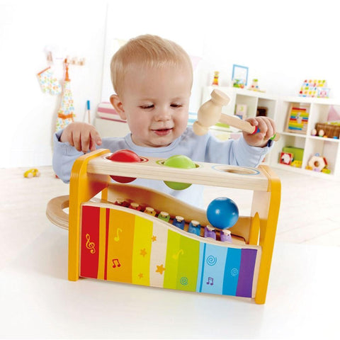 A baby plays with the Hape Pound and Tap Bench, a colorful wooden hammer toy from Hape. The toy features three balls that the baby hits with a mallet, causing them to roll down and strike metal xylophone bars, producing musical notes. This vibrant musical gift boasts a rainbow pattern and musical symbols, perfect for little ones.