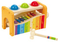 The Hape Pound and Tap Bench by Hape is a vibrant wooden hammer toy, perfect as a musical gift. It features three balls in blue, green, and red on top that can be struck with a mallet. Below, there is a metal xylophone with rainbow-colored keys and musical symbols. The mallet has a white handle decorated with red, blue, yellow, and green stripes.