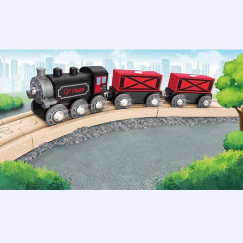 Illustration of the Hape Steam-Era Freight Train set crossing a wooden bridge. The classic train toy features a black engine with the word "Hape" and a red heart on the side, pulling two red cargo cars. Green trees frame the scene, with a city skyline and blue sky in the background.