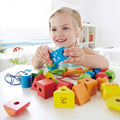 A young girl smiles as she plays with her colorful Hape String-Along Shapes on a white table. The wooden geometric blocks, offered by the brand Hape, come in various shapes, including cylinders, cubes, and triangles with different designs on them. This engaging activity enhances her fine motor development amidst the room's bright, clean, and minimalistic decor.