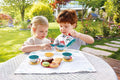 Two children, a blonde-haired girl and a red-haired boy, are engaging in pretend play at a table in the garden. They have the Hape Tea Time Playset by HAPE and toy snacks arranged on a white cloth. The background features lush greenery, flowers, and a pathway leading to a house.