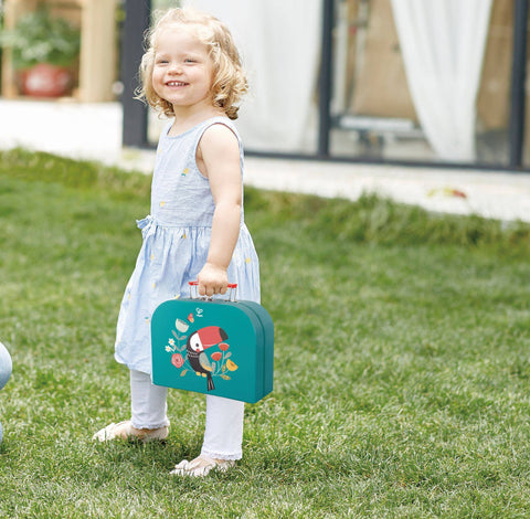 A young child with blonde curly hair wears a light-blue dress and white leggings, standing on grass and smiling. The child holds the Hape Tea Time Playset, a teal-colored lunchbox decorated with an illustration of a bird, perfect for pretend play. In the background, there is a building with a curtain.