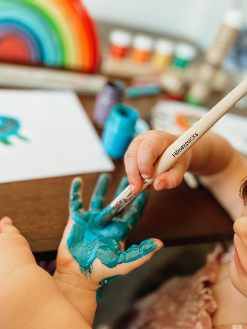 A child uses a Honeysticks Jumbo Paint Brush Set - 3 pack to paint their hand blue with a brush labeled "Honeysticks." On the table, there are various non-toxic paint bottles, a piece of paper with some artwork, and a colorful rainbow decoration in the background.