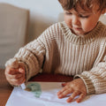 A young child with brown hair, dressed in a beige knitted sweater, is using a Honeysticks My First Paint Brush from the Honeysticks My First Paint Brush Set - 3 pack, equipped with an ergonomically designed handle, to create a watercolor painting on a white piece of paper. The child is focused and appears to be enjoying the activity.