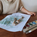 A child wearing a beige sweater is seated at a wooden table, painting. Two of the Honeysticks My First Paint Brushes from the Honeysticks My First Paint Brush Set - 3 pack lie on a partially painted white sheet of paper, showing green and blue hues. In the foreground is a metal watercolor paint palette with an assortment of colors.