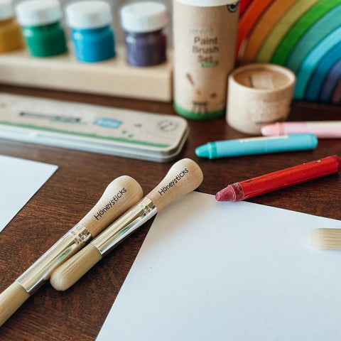 A close-up of an art workspace featuring various art supplies on a wooden table. Prominently displayed are paintbrushes from the "Honeysticks My First Paint Brush Set - 3 pack" with ergonomically designed handles. Surrounding them are crayons, jars of paint, a wooden box of colored pencils, and a rainbow-colored stacking toy in the background. White paper sheets are also visible.