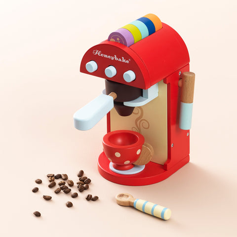 A vibrantly painted Le Toy Van Cafe Machine toy coffee maker set by LE TOY VAN features the word "Honeybake." It comes with a red coffee cup adorned with white polka dots, a wooden spoon, colorful pods on top, and scattered coffee beans on a light beige surface.