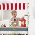 A child plays with the Le Toy Van Cafe Machine by LE TOY VAN, using a brightly painted drinks machine under a red and white striped awning. The stand includes various toy coffee cups, colorful pods, and accessories. The child is wearing a beige striped shirt and holding a toy scoop.