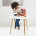 A young child with curly hair is playing with the Le Toy Van Cafe Machine, a brightly painted drinks machine by LE TOY VAN, on a small white table with wooden legs. The child is wearing a cream-colored top with colorful prints and blue pants, pretending to make coffee using colorful pods and accessories like cups and beans on the table.