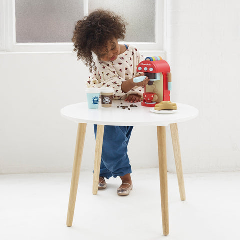 A young child with curly hair is playing with the Le Toy Van Cafe Machine, a brightly painted drinks machine by LE TOY VAN, on a small white table with wooden legs. The child is wearing a cream-colored top with colorful prints and blue pants, pretending to make coffee using colorful pods and accessories like cups and beans on the table.