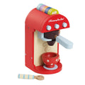 The Le Toy Van Cafe Machine by LE TOY VAN is vividly painted in red with a white and beige panel, featuring three buttons, a handle, and a cup holder. A red toy cup with white polka dots sits on the holder. A wooden spoon and colorful pods are placed beside the machine, while the brand name "Honeybake" is visible on top.