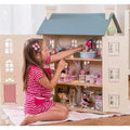 A young girl in a pink striped dress plays with a large LE TOY VAN Cherry Tree Hall dollhouse. The four-storey Le Toy Van Cherry Tree Hall, styled dollhouse, is open to reveal multiple rooms furnished with miniature furniture and dolls. She is sitting on a white rug arranging items inside the dollhouse.