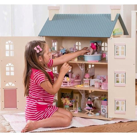 A young girl in a pink striped dress plays with a large LE TOY VAN Cherry Tree Hall dollhouse. The four-storey Le Toy Van Cherry Tree Hall, styled dollhouse, is open to reveal multiple rooms furnished with miniature furniture and dolls. She is sitting on a white rug arranging items inside the dollhouse.