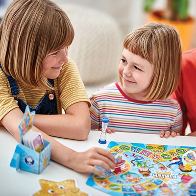 Two young children are sitting at a table playing the Orchard Toys What a Performance Game by Orchard Toys. They are smiling and looking at each other. The game includes activity cards, a sand timer, and a performance board with various paths and pictures. The background is a cozy indoor setting.