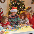 A family of four gathers around the table, delighting in a game of Orchard Toys What a Performance from Orchard Toys. Two children and an adult are beaming with joy as the third child, donning a Santa hat and festive sweater, examines an activity card. A decorated Christmas tree in the background enhances the holiday ambiance.