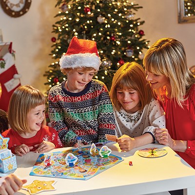 A family of four gathers around the table, delighting in a game of Orchard Toys What a Performance from Orchard Toys. Two children and an adult are beaming with joy as the third child, donning a Santa hat and festive sweater, examines an activity card. A decorated Christmas tree in the background enhances the holiday ambiance.