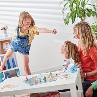 A young girl energetically stands and gestures with a raised arm, resembling a hero, while two seated individuals, likely her family members, watch and smile. They are in a bright room with *Orchard Toys What a Performance Game* by *Orchard Toys* on the table and a potted plant in the background.