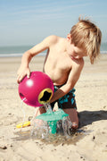 A young boy, sporting blue swim trunks adorned with a floral pattern, is playing on the beach. He is pouring water out of a QUUT Ballo Bucket in green, which is BPA-free and designed in the shape of a ball with a spout, onto the sand. The background displays a sandy beach and the ocean under a clear blue sky.