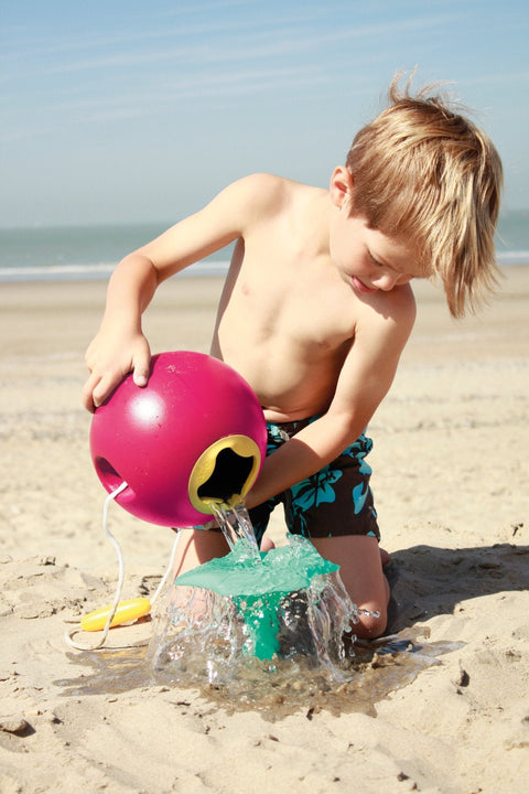 A young boy, sporting blue swim trunks adorned with a floral pattern, is playing on the beach. He is pouring water out of a QUUT Ballo Bucket in green, which is BPA-free and designed in the shape of a ball with a spout, onto the sand. The background displays a sandy beach and the ocean under a clear blue sky.