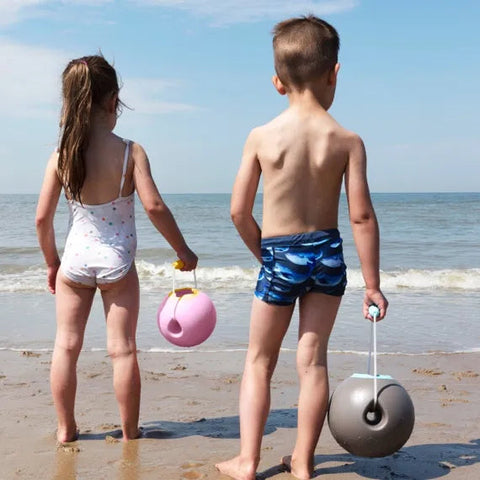 Two children, one in a white swimsuit and the other in blue swim trunks, stand on a sandy beach facing the ocean. Each child is holding a QUUT Mini Ballo Bucket - Banana Blue with soft-touch finishing—one pink and one gray—as they look out towards the water on a sunny day.