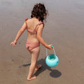 A young child with curly hair, wearing a pinkish swimsuit, is walking on a sandy beach. They are carrying a QUUT Mini Ballo Bucket in Banana Blue with smart design and soft-touch finishing in their right hand. The beach is wet, suggesting proximity to the water. The child is looking down and facing away from the camera.