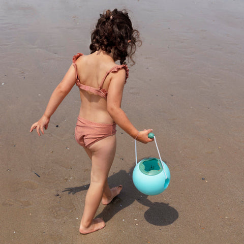 A young child with curly hair, wearing a pinkish swimsuit, is walking on a sandy beach. They are carrying a QUUT Mini Ballo Bucket in Banana Blue with smart design and soft-touch finishing in their right hand. The beach is wet, suggesting proximity to the water. The child is looking down and facing away from the camera.
