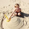 A young boy with blonde hair sits on the sandy beach, smiling brightly at the camera. He is shirtless and wears patterned swim trunks. Using the QUUT Triplet Beach & Bath Toy in Cherry Red from QUUT, he shapes a sand sculpture with precision. The background shows the smooth, undisturbed sand of the beach.