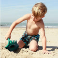 A young boy with light hair and wearing patterned swim trunks is playing with the QUUT Triplet Beach & Bath Toy in Cherry Red on a sandy beach. The sea and clear sky are visible in the background.