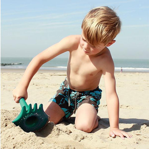 A young boy with light hair and wearing patterned swim trunks is playing with the QUUT Triplet Beach & Bath Toy in Cherry Red on a sandy beach. The sea and clear sky are visible in the background.