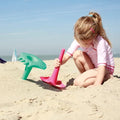 A young child with blonde hair, tied in a ponytail, is playing in the sand at the beach. The child is using colorful, multifunctional beach toys from QUUT, including the red QUUT Triplet Beach & Bath Toy - Cherry Red, to dig in the sand. The sky is clear and blue, and the scene appears sunny and bright.