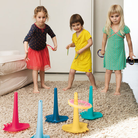 Three young children are playing an indoor ring toss game on a beige carpet. They are using QUUT Triplet Beach & Bath Toys in Cherry Red, which are placed on the floor and resemble beach toys. Each child is holding rings, aiming to toss them onto the QUUT cones. The children are dressed in various colorful outfits and appear focused on the game.