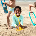 Three children are playing on a sandy beach, digging and building with bright multifunctional sand tools. The child in the center, wearing a light blue long-sleeved shirt, smiles broadly with their tongue out while holding a QUUT Triplet Beach & Bath Toy - Cherry Red shovel.