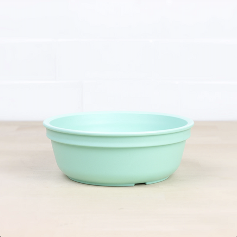 The Re-Play Bowl by REPLAY, a vibrant piece of tableware in light green, rests on a light wooden surface against a white tiled background. Featuring slightly tapered sides and a smooth finish, this round plastic bowl is made from recycled materials, making it safe for kids and perfect for any meal.