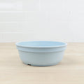 A Re-Play Bowl from REPLAY, made of light blue recycled plastic, sits empty on a light wooden countertop. The background features white tiled walls.