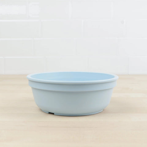 A Re-Play Bowl from REPLAY, made of light blue recycled plastic, sits empty on a light wooden countertop. The background features white tiled walls.