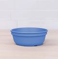 A Re-Play Bowl from our vibrant tableware set, produced by REPLAY, is placed on a light wooden surface against a white tiled background. Made from recycled plastic, the blue bowl features a simple design with a slight lip around the top edge and a smooth finish, ensuring it is safe for kids.