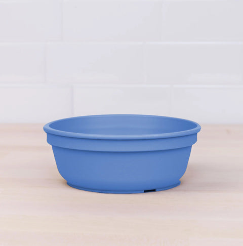 A Re-Play Bowl from our vibrant tableware set, produced by REPLAY, is placed on a light wooden surface against a white tiled background. Made from recycled plastic, the blue bowl features a simple design with a slight lip around the top edge and a smooth finish, ensuring it is safe for kids.